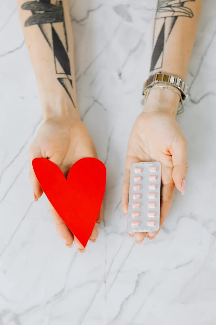Tattooed hands holding a red heart and blister pack of pills on marble surface.